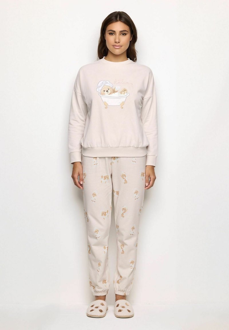 Mujer vestida con pijamas beige con ilustraciones de ositos de peluche y zapatillas a juego, de pie contra un fondo blanco.