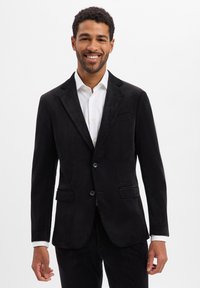 Blazer in suede nero con rever a incavo, due bottoni e tasche anteriori. Indossato sopra una camicia bianca con bottoni, tessuto liscio, vestibilità sartoriale.