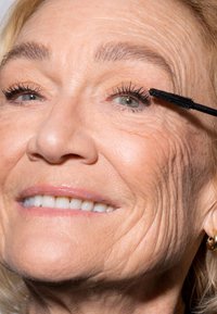 Svart mascara er påført krøllete øyevipper; huden viser naturlig tekstur og rynker. Subjektets lepper er lett glanset og smilende.