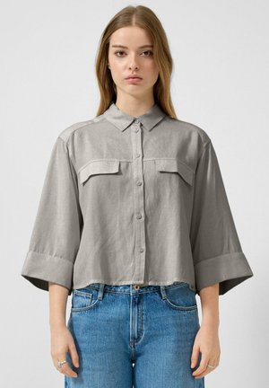 Młoda kobieta w luźnej, jasnoszarej koszuli na guziki o szerokich rękawach oraz niebieskich, wysokich dżinsach, stojąca na tle o jednolitym kolorze.
