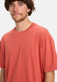 Jonge man met kort bruin haar, gekleed in een eenvoudig koraalrode T-shirt met korte mouwen en een subtiele geborduurde logo op de borst, neutrale achtergrond.