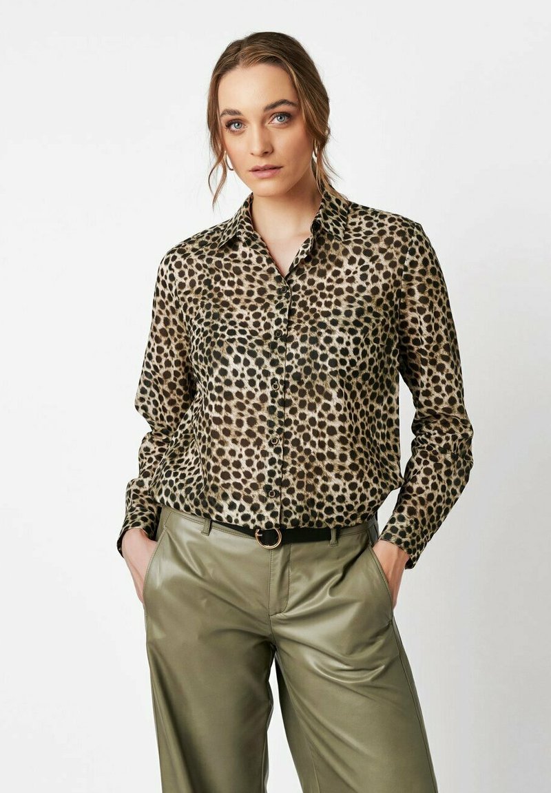 Woman wearing a leopard print button-up blouse tucked into high-waisted olive green leather pants with hands in pockets.