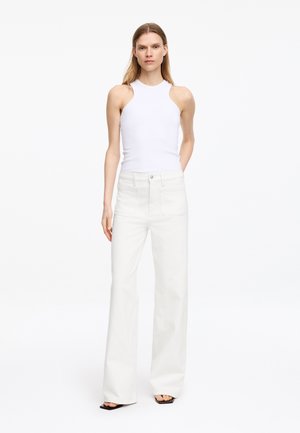 Frau steht und trägt ein ärmelloses weißes Neckholder-Top, hoch taillierte weite weiße Hosen und schwarze offene Sandalen vor einem weißen Hintergrund.