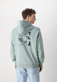 Felpa in cotone verde chiaro con cappuccio, caratterizzata da una grafica nera sul retro. Morbido al tatto, vestibilità rilassata e polsini a costine.