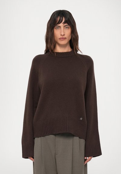 Femme avec des cheveux raides et foncés portant un pull oversize marron foncé et un pantalon large gris sur un fond clair uni.
