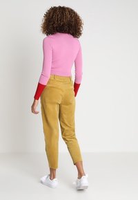 Eng anliegenden rosa Pullover mit roten Ärmeln, kombiniert mit gelben kordeligen Hosen in Knöchellänge. Modell trägt weiße Turnschuhe, Ansicht von hinten.