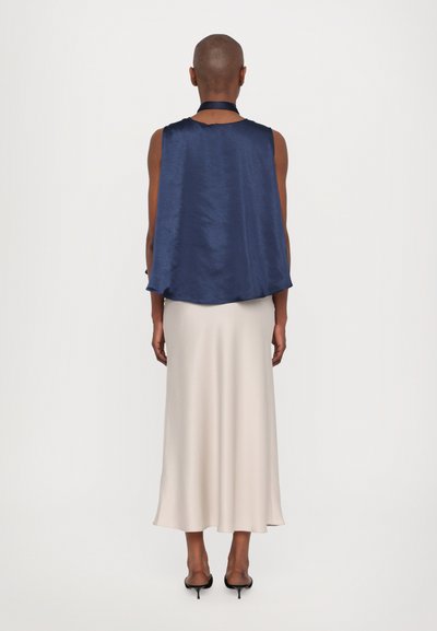 Navy satin sleeveless top with flowing design, paired with a light beige knee-length skirt. Model wears black open-toe heels.