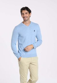 Pull bleu clair en V en maille, avec des manches longues et un petit logo noir sur la poitrine gauche, associé à un pantalon beige.