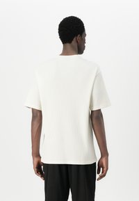 Fehér, texturált rövid ujjú, kerek nyakú póló. Laza szabású, oldalsó hasítékokkal, hátulról bemutatva.