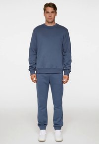 Miesten sininen collegepaita ja siihen sopivat collegehousut, molemmat pehmeästä kankaasta ja resorireunuksilla. Valkoiset lenkkarit täydentävät asun.