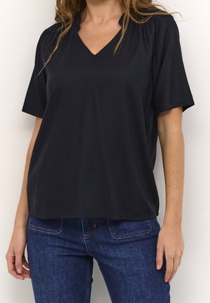 Femme portant un chemisier noir à manches courtes en V ample, associé à un jean denim bleu foncé, debout contre un mur blanc.