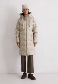 Nainen, joka on pukeutunut beigeseen puffer-takkiin, jossa on huppu ja pipot, yhdessä ruskeiden leveälahkeisten housujen ja tummanruskeiden loafereiden kanssa, seisoo sisätiloissa.