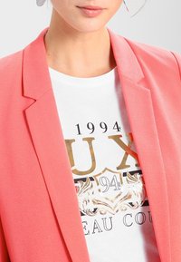 Blazer corail à la coupe ajustée, associé à un t-shirt blanc graphique orné de motifs et embellissements dorés et noirs complexes.