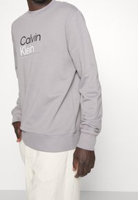 Felpa grigia con logo "Calvin Klein" nero, design con collo rotondo, polsini e orlo a coste, con una finitura in tessuto morbido e strutturato.