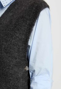 Gilet di lana grigia e morbida con scollatura a V profonda, indossato sopra una camicia blu chiaro con bottoni, caratterizzato da un bottone decorativo in argento sul lato.