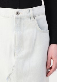 Jupe en denim bleu clair à taille haute, avec une poche avant et un bouton en métal argenté. Le tissu a une texture douce et un léger dégradé.