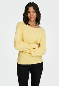 Jasnożółty prążkowany sweter z szerokim dekoltem i długimi rękawami, zestawiony z czarnymi spodniami. Miękka faktura z delikatnymi pionowymi paskami.