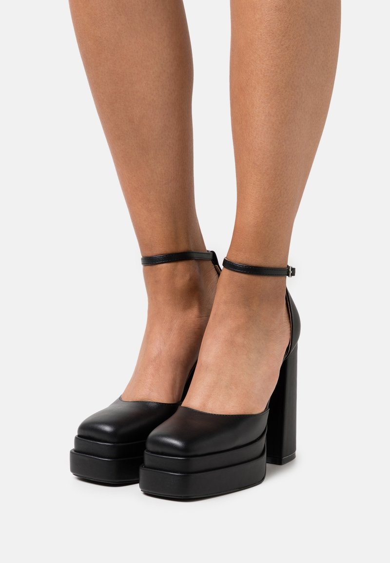 Jambes portant des talons hauts à plateforme noirs avec des brides à la cheville, debout sur un fond blanc.