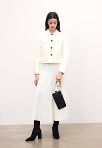 Vit kort jacka med svarta knappar, kombinerad med en lång vit kjol, svarta stövlar med klack och en liten svart handväska med kedjerem.