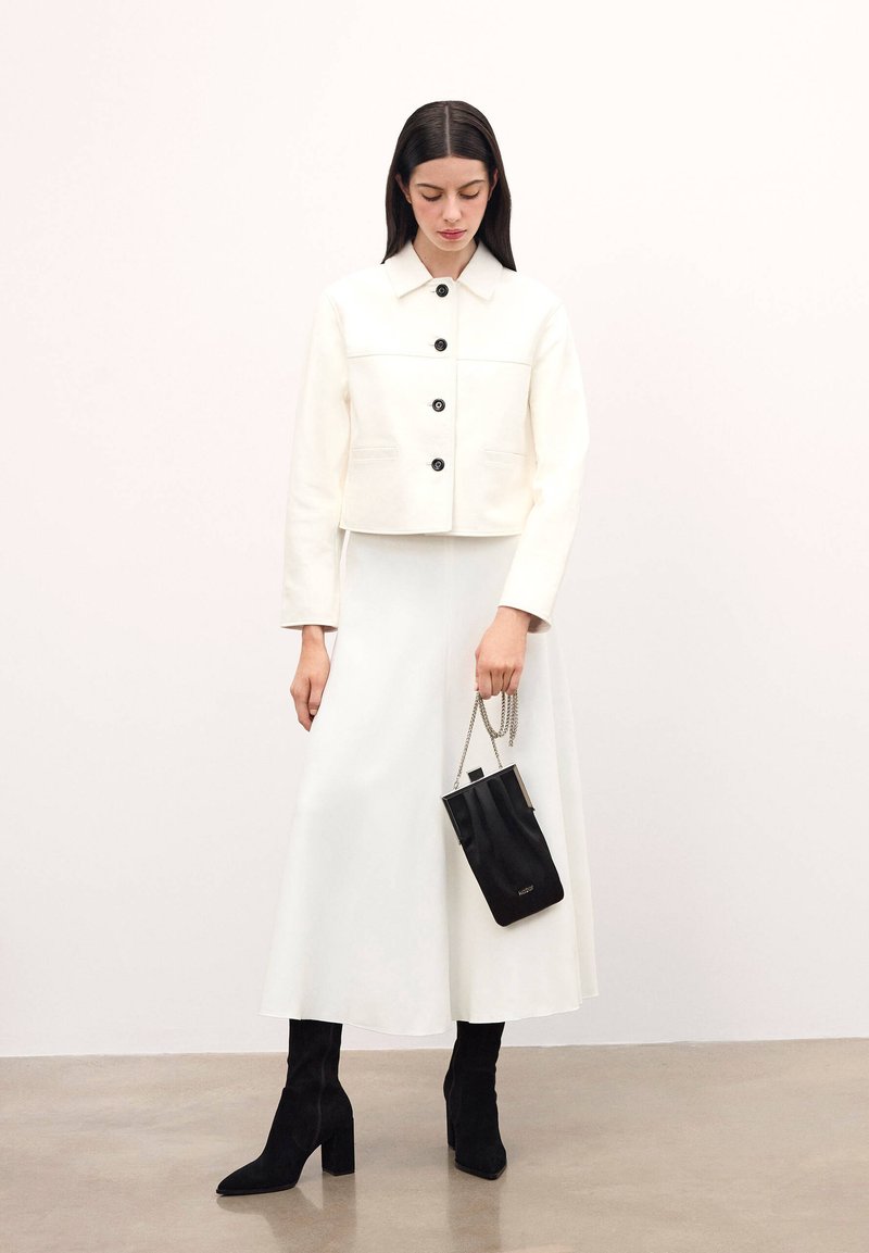 Vit kort jacka med svarta knappar, kombinerad med en lång vit kjol, svarta stövlar med klack och en liten svart handväska med kedjerem.