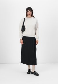Licht beige sweatshirt met tekst, zwarte midi-rock, zwarte loafers en een kleine zwarte schoudertas. Model staat tegen een witte achtergrond.