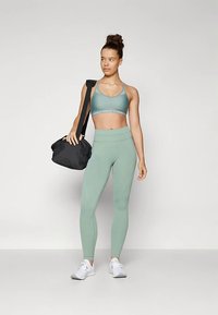 Αθλητική εμφάνιση με μπλουζάκι σπορ σε τιρκουάζ και αντιστοιχικά ψηλόμεσα κολάν. Μια μαύρη τσάντα duffel κρέμεται από τον ώμο. Λευκά αθλητικά παπούτσια.