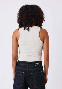 White ribbed tank top viewed from the back, paired with dark denim pants featuring a branded patch on the waistband and contrasting stitching.
