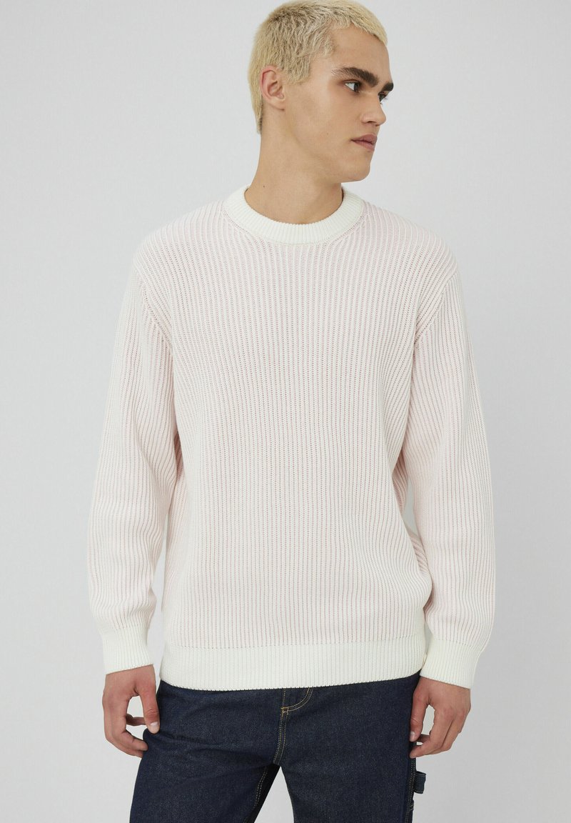 Pull côtelé rose clair avec bord blanc. Caractéristiques : manches longues et col rond. Tissu texturé avec une finition douce, associé à un jean foncé.