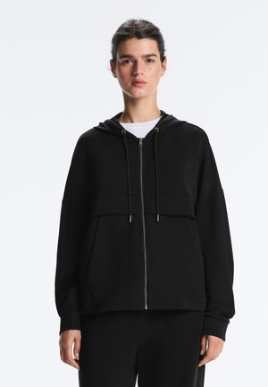 Nainen pukeutuneena mustaan vetoketjulliseen huppariin, jossa on etutaskut ja kiristysnyörit, valkoisen paidan päällä, seisoo yksinkertaisen vaalean taustan edessä.