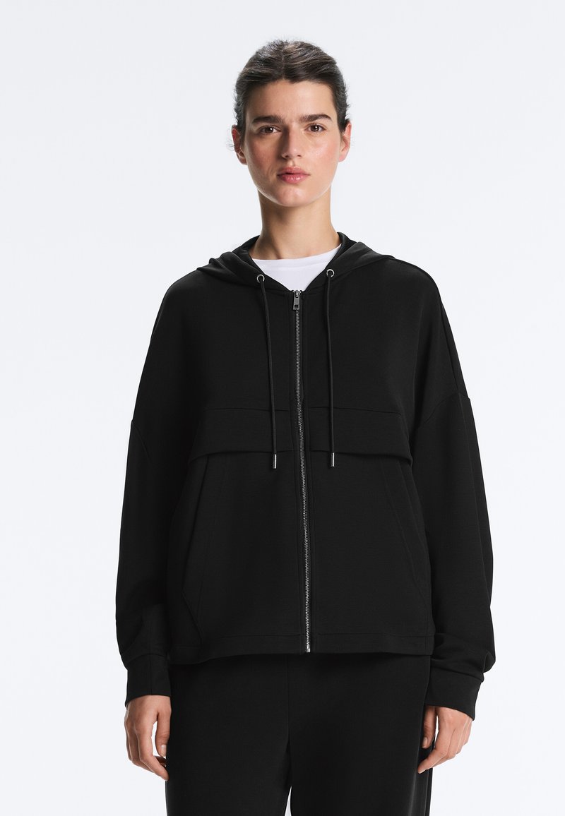 Femme portant un sweat à capuche noir zippé avec poches avant et cordons, par-dessus un t-shirt blanc, debout devant un fond clair uni.