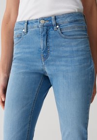 Jean en denim bleu clair avec une silhouette ajustée, présentant une fermeture à boutons classique, des passants de ceinture et des poches avant avec des détails de couture subtils.