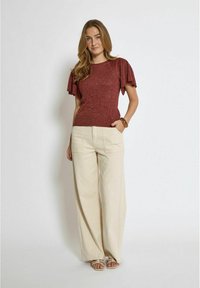 Red glitter top with flared sleeves paired with high-waisted cream wide-leg trousers. Model poses with one hand in pocket, wearing sandals.