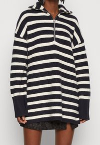 Sweter z paskami w kolorze granatowym i białym, z zamkiem błyskawicznym przy kołnierzu, prążkowaną fakturą, luźnym krojem i długimi rękawami.