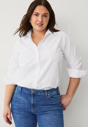 Chemise blanche à boutonnage, avec col et manches retroussées, associée à un jean en denim bleu présentant un léger délavage et des détails de poches avant.
