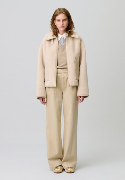 Giacca in felpa beige con colletto, maglione beige chiaro e pantaloni larghi a righe beige. Il modello indossa sneakers beige. Sfondo neutro.