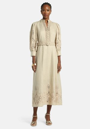 Kvinde iført en lang beige kjole med puffede ærmer og blonderudsnit, et matchende bælte og sandaler med stropper, stående mod en hvid baggrund.