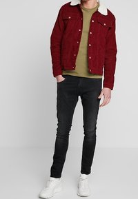 Rode corduroy jas met witte bontkleurige kraag, olijfgroene T-shirt, zwarte slim-fit jeans en witte sneakers. Casual outfit met gelaagde texturen.