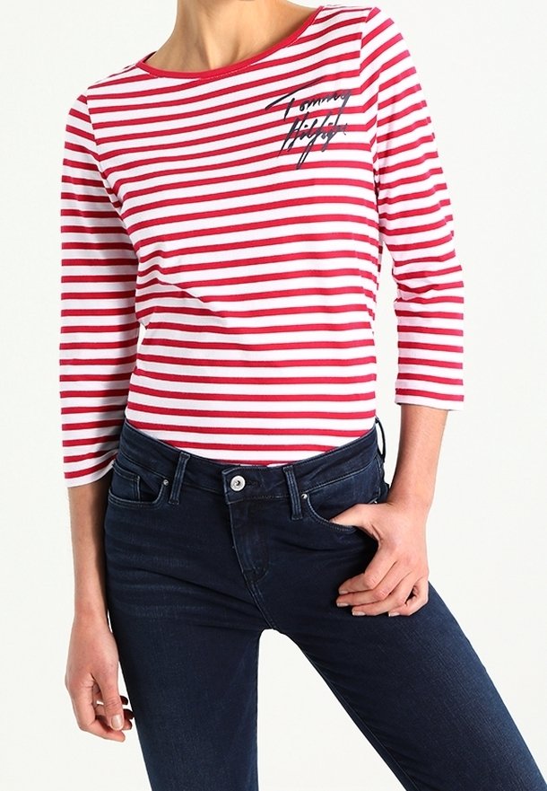 T-shirt à manches longues rayé rouge et blanc avec un décolleté bateau et un logo noir, associé à un jean bleu marine. Matière en coton lisse.