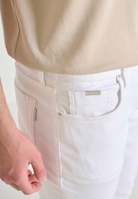 Jeans in denim bianco presentano una tasca posteriore con un'etichetta argentata. Una camicia beige è parzialmente visibile sullo sfondo. La texture appare liscia.