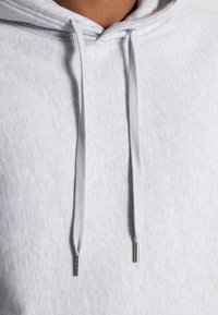 Světlě šedá mikina s kapucí a texturovaným povrchem. Má stahovací šňůrky se stříbrnými konci, prostornou kapuci a žebrovaný výstřih.