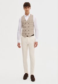 Beżowa kamizelka w kratę z podwójnymi guzikami, biała koszula, kremowe spodnie i brązowe buty. Prosty fason z dopasowanym krojem.