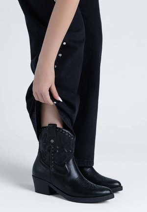 Persona que lleva pantalones negros con botones plateados, levantando la pernera para mostrar botas negras de cuero hasta el tobillo con costuras decorativas y tachuelas.