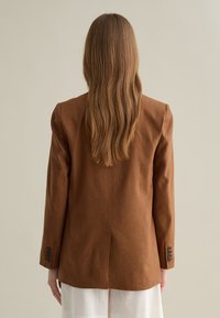 Blazer di lino marrone con vestibilità su misura, dotato di spalline, polsini bottone e spacco sul retro. Tessuto uniforme, dettagli minimal.