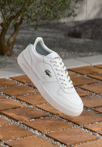 Witte leren sneaker met groene accenten, ronde neus, gestructureerde zool, geperforeerd ontwerp en vetersluiting, met een Lacoste-logo.