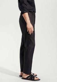 Man wearing black trousers and a black long-sleeve shirt, standing barefoot with black woven leather slide sandals against a plain background.