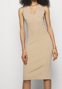 Femme portant une robe sans manches beige en maille avec un col en V, un collier chaîne doré et un bracelet doré sur un fond uni.