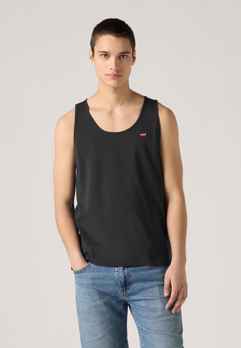 Νέος άνδρας με κοντά μαλλιά, φοράει μαύρο αμάνικο τοπ Levi's και ανοιχτό μπλε τζιν, στέκεται μπροστά σε απλό φωτεινό φόντο.