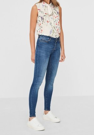 Kvinde iført ærmeløs blomstret bluse med bundet hals, blå skinny jeans og hvide sneakers, stående mod en ensfarvet lys baggrund.