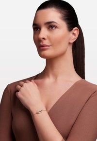 Sidabrinė apyrankė su susipynusiais auksiniais ir sidabriniais akcentais ant subtilios grandinėlės, dėvima ant nuogas riešo, prieš neutralią foną.
