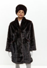 Brun faux fur frakke med en bred krave, to sidelommer og matchende pels hat. Teksturen er tyk og fyldig, hvilket skaber et varmt udseende.
